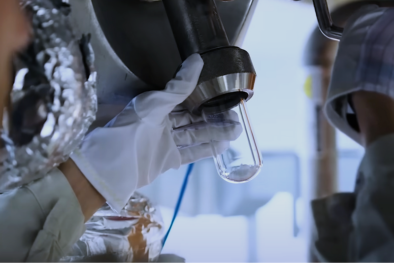 Person in a lab setting holding a glass container with a metallic device